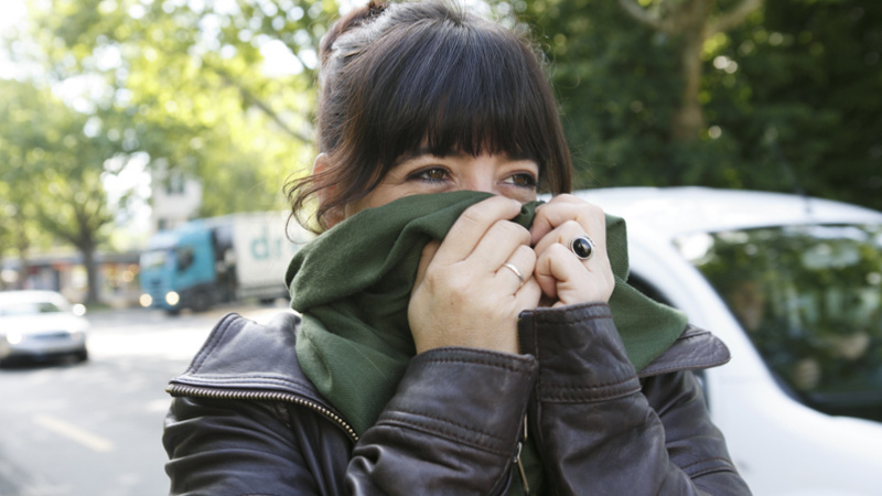 Heuschnupfen, Allergie, Pollenallergie: junge Frau in einer Stadt an einer vielbefahrenen Strasse, sie hat sich den grünen Schal über Mund und Nase gezogen