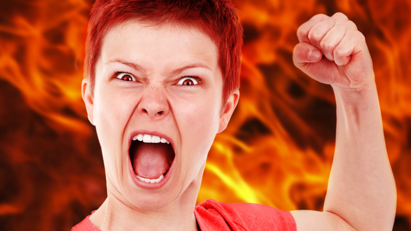 Psychologie, Aggression, ärgern: Frau mit kurzen roten Haaren hat die linke Hand zur Faust geballt und den Mund zum Schrei geöffnet. Im Hintergrund lodern Flammen