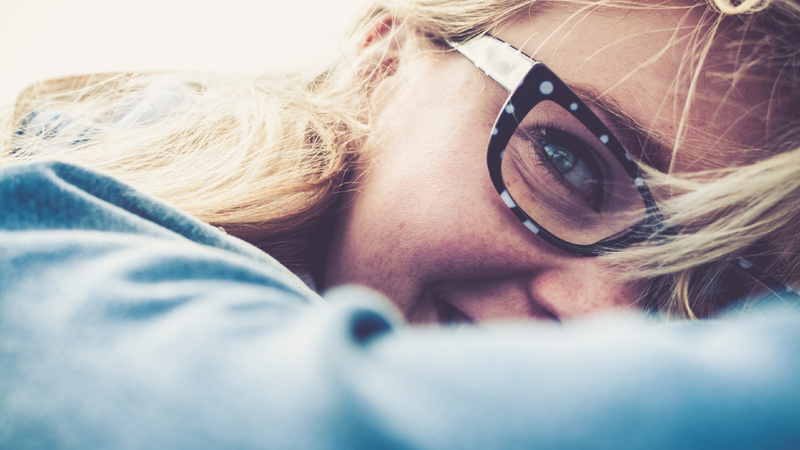 Akne, Pickel, unreine Haut: Jugendliche mit langen blonden Haaren und Brille, man sieht nur die Hälfte der rechten Gesichtshälfte. Sie hat den Kopf auf den Arm gebettet