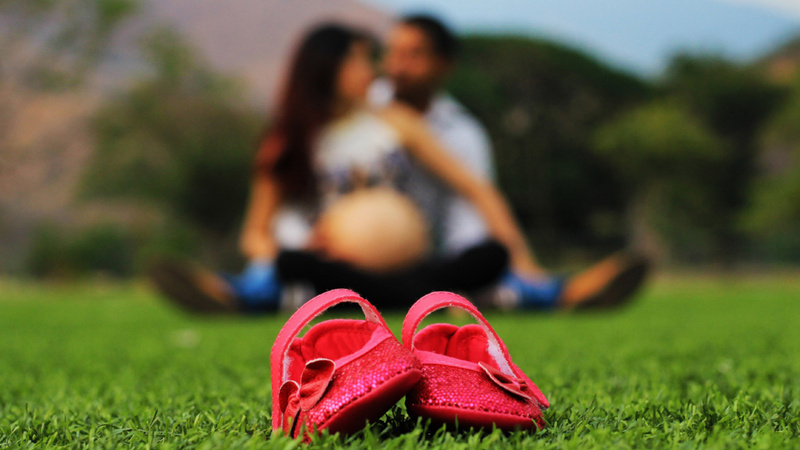 Ein Päärchen sitzt auf dem Rasen. Sie ist schwanger. Vor ihr liegt ein paar Babyschuhe.