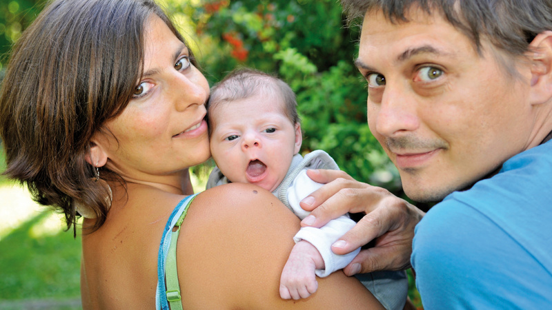 Sexualität, Geburt: Mutter und Vater mit ihrem Neugeborenen. Die Mutter hält das Kind im Arm, es schaut ihr über die rechte Schulter, der Vater steht daneben und hält das Baby am Arm fest