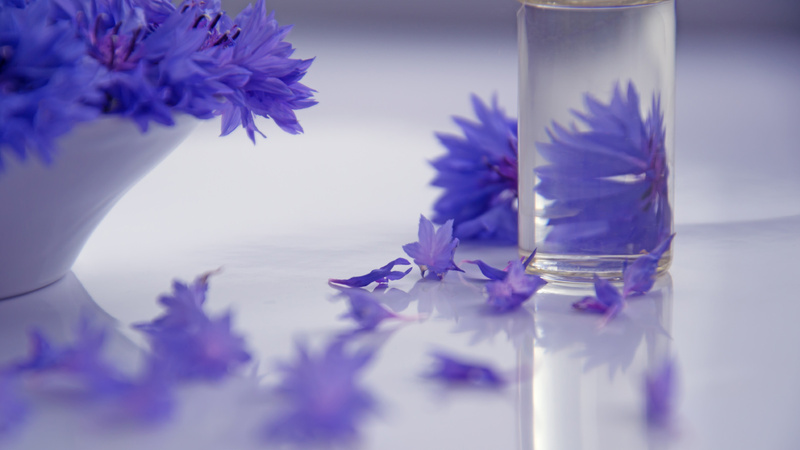 Ein durchsichtiges Fläschchen, darin eine klare Flüssigkeit. Im Hintergund durscheinend eine violette Blüte. Daneben steht eine Schale mit denselben violetten Blüten.