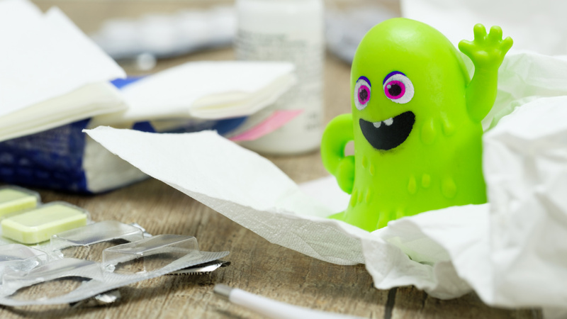 Erkältung, Viren, Forschung: Tisch mit allerlei Kram wie Papiertaschentücher, Tabletenblister etc. Darin ein grüner Plastikvirus, der ein hämisches Gesicht macht und winkt