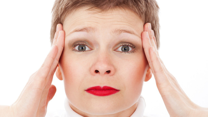 Kopfschmerzen, Kopfweh, Ursachen: Frau mit kurzen braunen Haaren hält sich beide Hände an die Schläfen, ihr Gesicht ist schmerzverzerrt