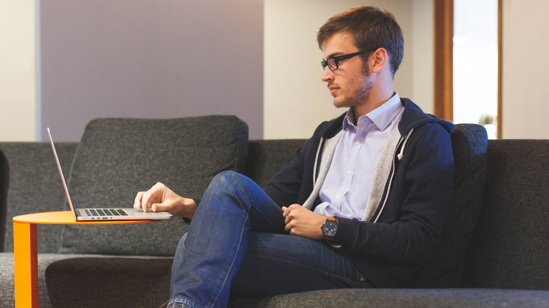 Un homme est assis sur un canapé et consulte un ordinateur portable devant lui.