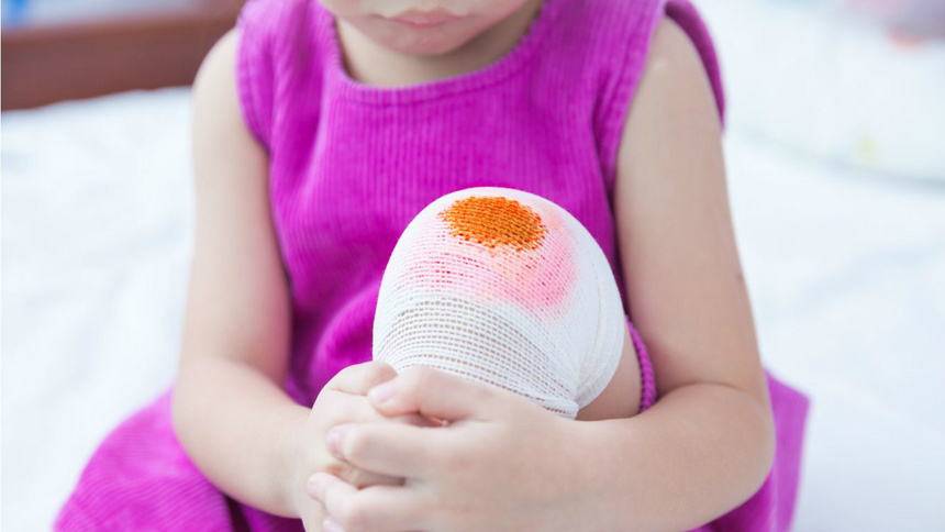 Une petite fille avec une robe rose qui a le genou dans dans un pansement.