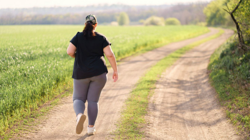 Fitness, Sport, Übergewicht: Man sieht eine übergewichtige Frau von hinten, die auf einem Weg in einem Feld joggt.
