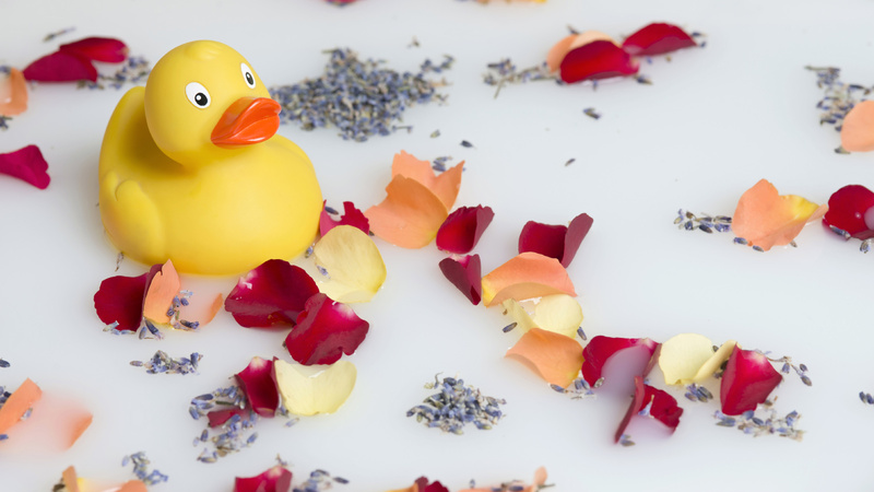 Bien-être bains huiles essentielles: un canard jaune de bain entouré de pétales et de grains de lavande.