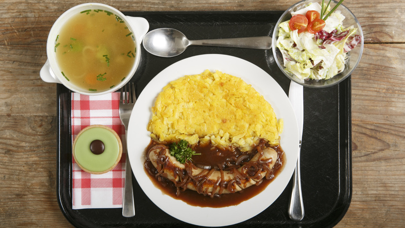 Ernährung, Mittagessen, gesund: Tablett mit einer klaren Gemüsesuppe in einem Schälchen, einem gemischten Salat in einem Schälchen, einem weissen Porzellanteller mit einer Bratwurst an Zwiebelsauce und Rösti, auf einer rot-weiss karierten Serviette liegt ein Carrac. Messer, Gabel und Löffel liegen parat.