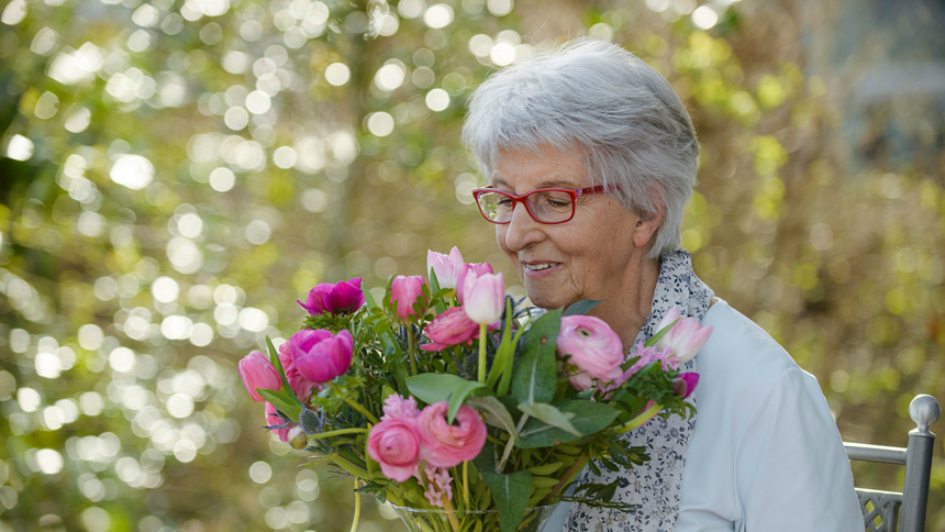 Sehen, riechen, hören: Eine alte Frau riecht an einem rosafarbenen Blumenstrauss.