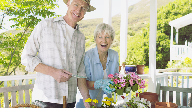 Schönheit, Pflege, Haare, grau: Mann und Frau auf einer Veranda, er trägt einen Strohhut. Sie beide pflanzen Blumen in Töpfe. Die grauhaarige Frau lacht