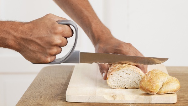 Zwei Hände, die eine hält ein Brot fest, das auf einem Brotbrett aus Holz liegt. Die andere hält ein Brotmesser mit speziellem Griff und schneidet gerade ein Stück ab