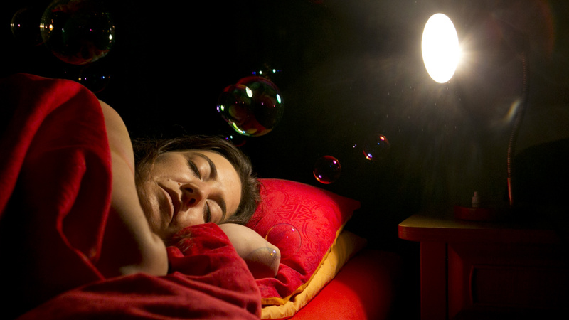 Schlafen, Träume, Träumen: Frau liegt in einem Bett mit roter Bettwäsche auf der linken Seite. Über ihr schweben Seifenblasen, oben rechts leuchtet ein Licht.