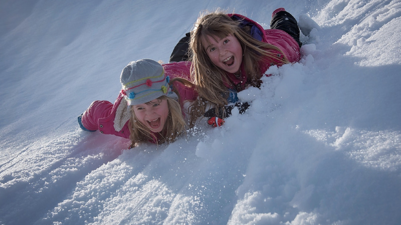 Winter, Schnee, Kälte: Zwei Mädchen rutschen bäuchlings einen verschneiten Hang hinunter und amüsieren sich sehr.