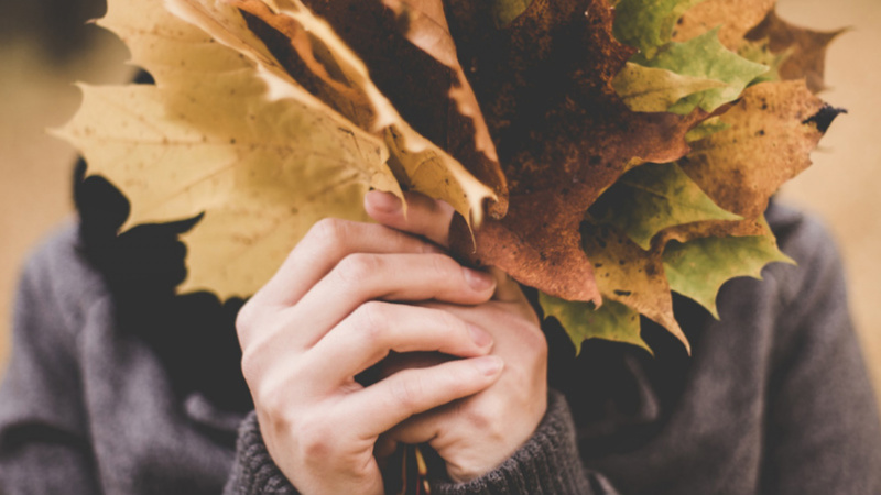 Une femme se cache derrière un bouquet de feuilles mortes. On ne voit que ses mains.
