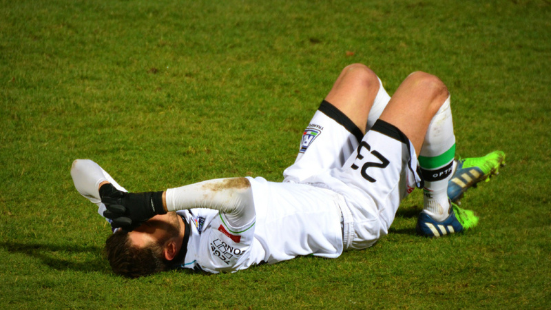 Sportverletzungen, Verstauchung, Prellung: Fusssballer in weissem Fussballdress liegt rücklings auf dem Rasen, die Beine angewinkelt, die hände an den Kopf gelegt.
