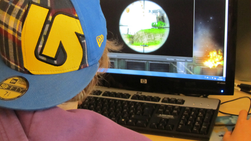 Sucht, Gamesucht, Computer: Ein Kind mit verkehrt herum aufgesetzter Baseballmütze sitzt vor einem Computerbildschirm und spielt ein Spiel.