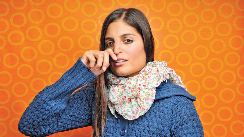 Junge Frau mit langen graden schwarzen Haaren in einem blauem Wollpullover reibt sich mit dem Zeigefinger unter der Nase durch