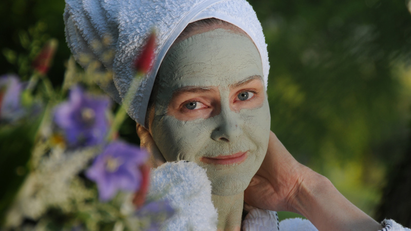Schönheit, Pflege, Gesicht, Gesichtsmasken: Frau mit weissem Frotteetuch um den Kopf gebunden hat eine grüne Gesichtsmaske auf dem Gesicht, unscharf im Vordergrund ein Blumenstrauss