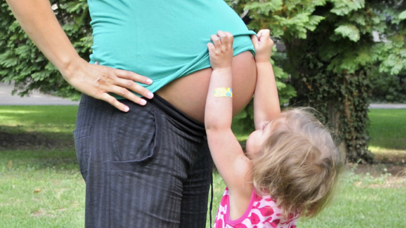 Une petite fille lève le pull de sa maman bien enceinte plantée devant elle pour regarder son ventre.