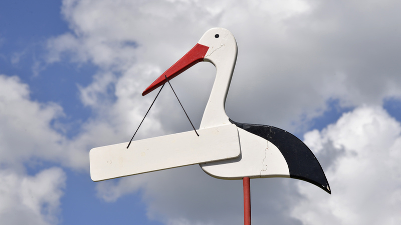 Schwangerschaft, Geburt, Name, Vorname: Ein Storch aus Holz der ein Schild im Schnabel trägt, das Schild ist weiss.