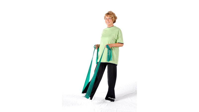 Alte Frau, gürnes Shirt, schwarze Hose, hat das rechte Bein in das Gummiband gesteckt und zieht mit beiden Händen am Band