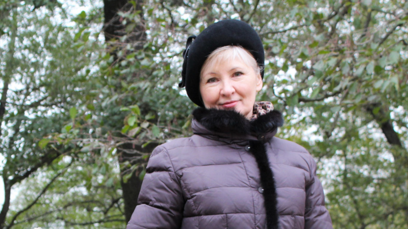 Beauté soins peaux matures: une femme d'un certain âge de face dans une forêt. Elle porte un bérêt noir et une doudoune violette.