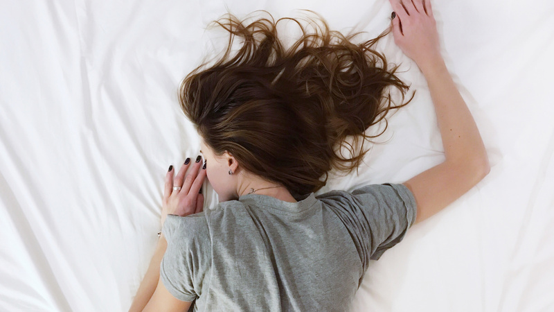 Schlafen, Regeneration, Nachtruhe: Frau liegt bäuchlings auf einem weiss bezogenen Bett, wir sehen nur den Oberkörper. Sie trägt ein graues T-Shirt, ihre langen braunen Haare liegen ausgebreitet neben ihrem Kopf.