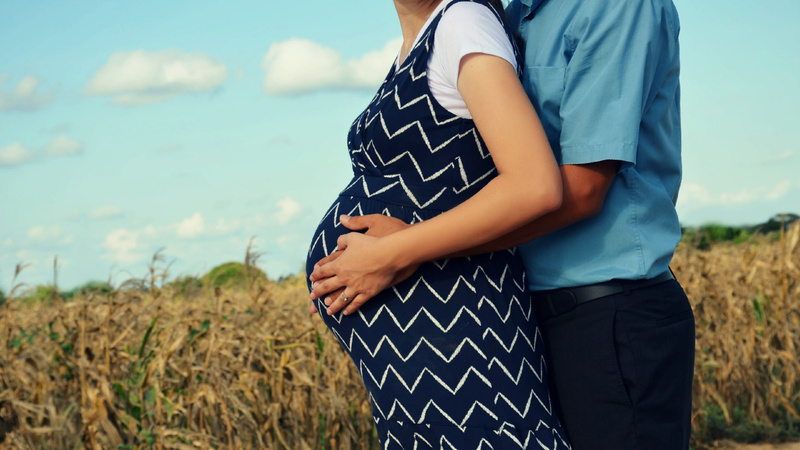 Ernährung, Mirkonährstoffe: Schwangere Frau und ein Mann. Er umfasst ihren Bauch von hinten mit den Händen. Sie stehen in einem Kornfeld.