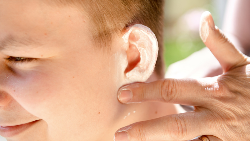 Une main de femme applique de la crème solaire sur l'oreille d'un garçon.