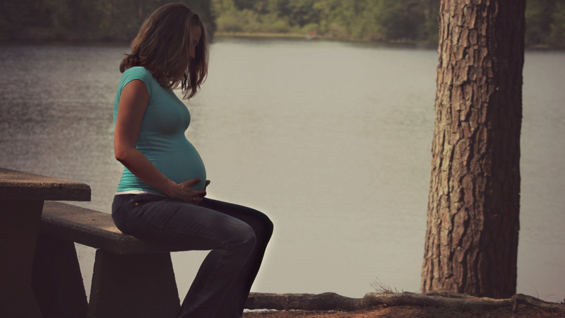 Schwangerschaft, Schwangerschaftsbeschwerden, Übelkeit: Schwangere sitzt auf einer Bank an einem See und hält sich den BAuch