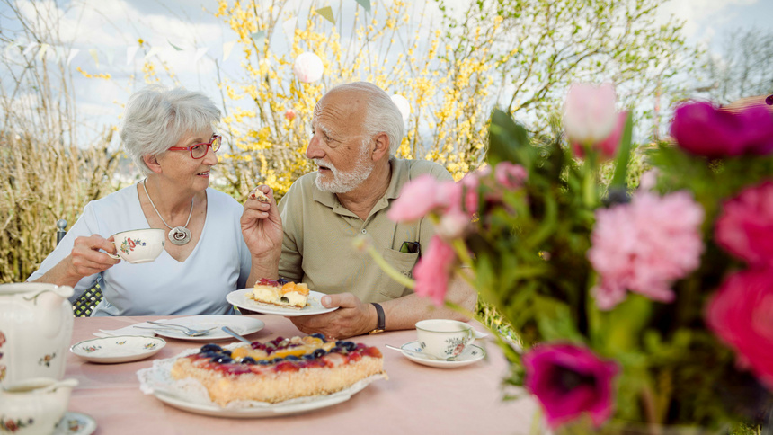 Geschmack, Geschmackssinn, Ageusie: eine alte Frau und ein alter Mann sitzen an einem Tisch im Freien. Auf dem Tisch steht ene Früchtetorte. Der Mann hält einen Teller mit einem Stück Torte in der Hand, die Frau eien Teetasse. Im Vordergrund ist unscharf ein Blumenstrauss zu sehen.
