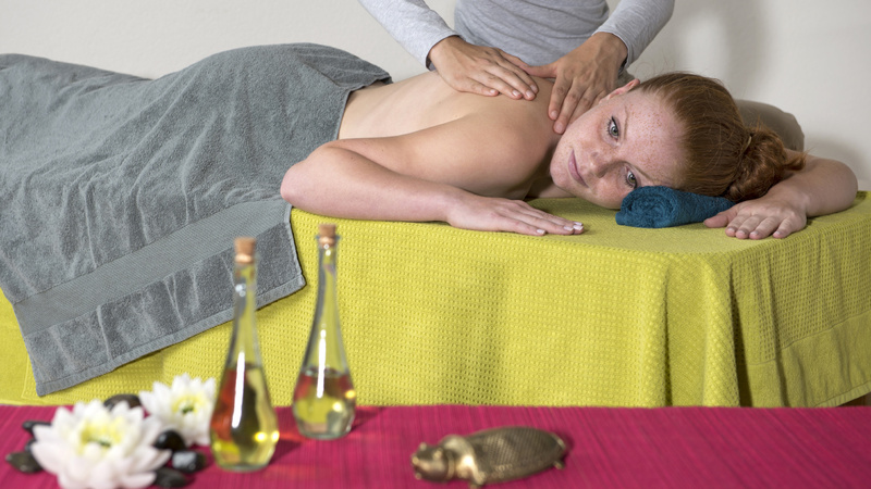 Une femme est couchée sur une table de massage et une autre femme la masse. Devant, il y a des fioles d'huiles essentielles.
