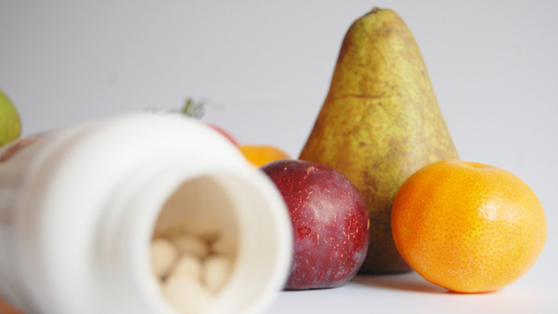 Mineralstoffe, Vitamine, Vitalstoffe, Immunsystem: Plastikdöschen liegt geöffnet auf der Seite, darin Vitaminpillen. Im Hintergrund eine Birne, eine Mandarine und eine Pflaume