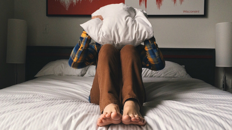 Ein Mann sitzt auf einem Bett und hält ein Kissen vor sein Gesicht.