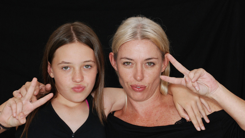 Pubertät, Eltern, Kinder, Konflikt: Tochter und Mutter stehen nebeinander, beide tragen schwarze Oberteile. Sie haben sich gegenseitig den Arm um die Schulter gelegt udn machen beide das Victory-Zeichen mit der Hand.