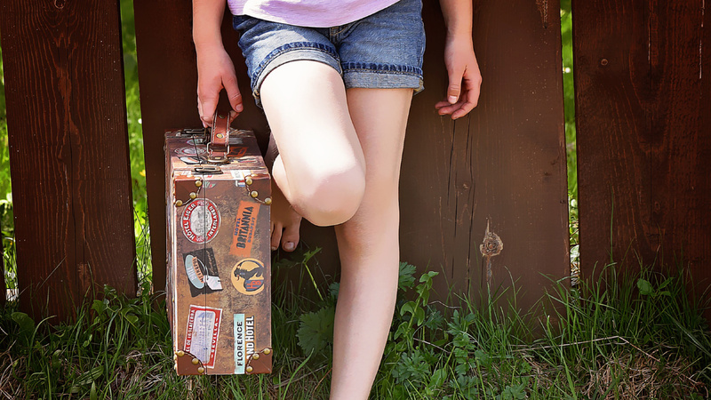 Pubertät, Reisen, Urlaub, Ferien: Junge Frau mit kurzen blauen jeans, wir sehen nur die Beine. Sie hält einen braunen Lederkoffer in der rechten Hand, der voller Aufkleber ist.