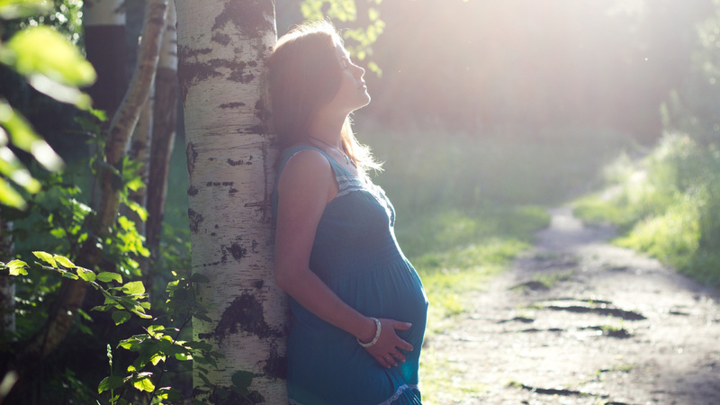 Conseils protection solaire femmes enceintes: une femme enceinte est appuyée contre un tronc d'arbre sur un chemin forestier. Elle tient son ventre.