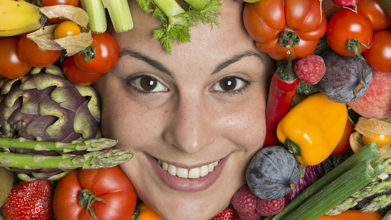 Ernährung, Vegetarismus, Risiken: Frauengesicht, das aus einem grossen Haufen Früchte und Gemüse herausschaut
