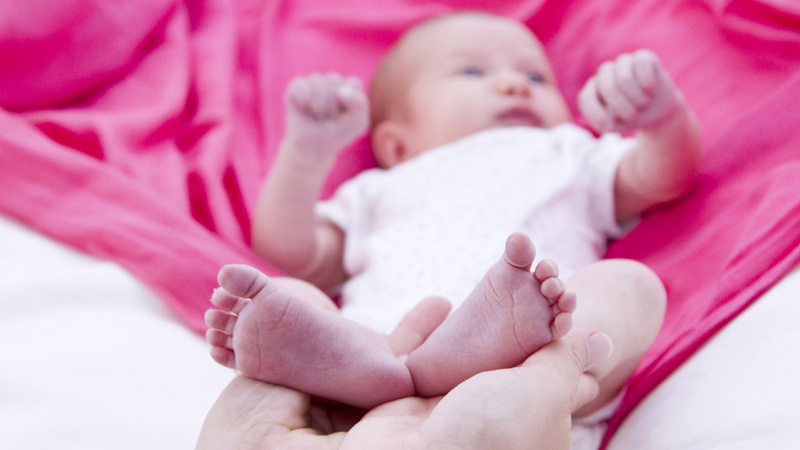 Schwangerschaft, Geburt, Stillen, Stilldemenz: Neugeborenes in weissem Gewand liegt auf einer rosafarbenen Decke, die Mutter hält es an beiden Füssen fest.