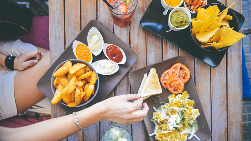 Alimentation, quantité, excès, carences: une table vue du dessus. On voit le bras d'une femme et de la nourriture de fast-food sur des assiettes. 