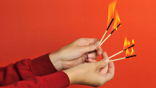 Deux mains tiennent des allumettes allumées sur un fond rouge.